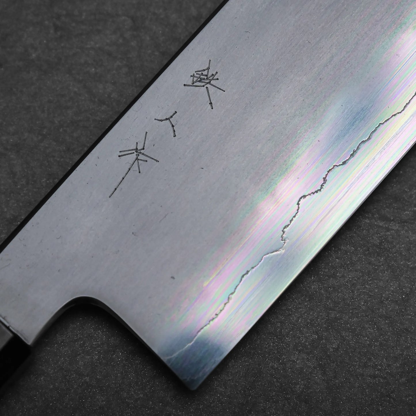 Close-up of a Japanese vegetable knife blade with engraved kanji characters and a visible wavy temper line, resting on a dark textured surface.