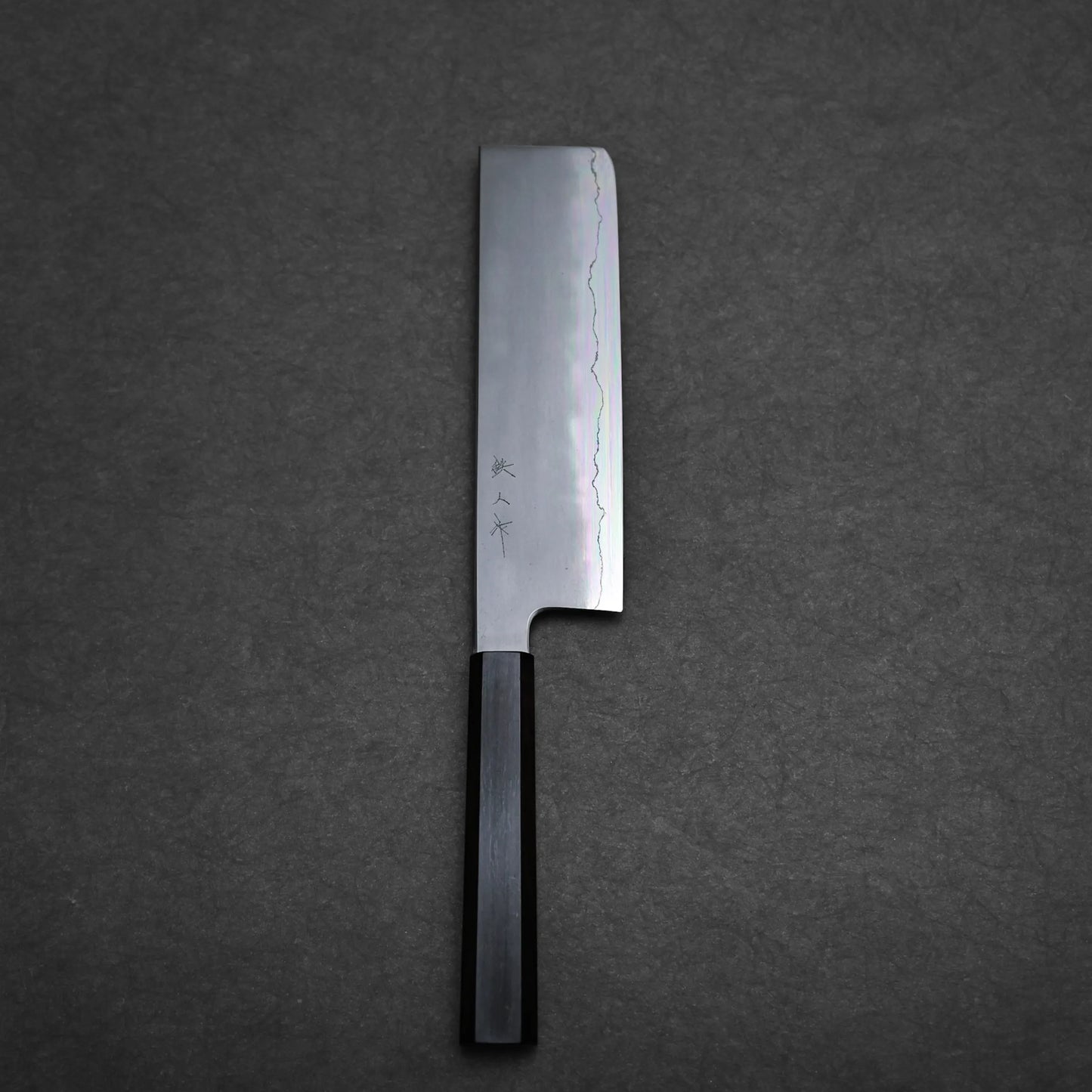 A Japanese vegetable knife with a rectangular blade and dark wooden handle rests on a dark textured surface. This nakiri knife features a wavy hamon pattern and Japanese characters near the handle.