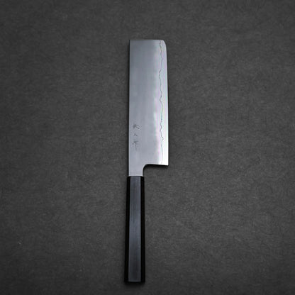 A Japanese vegetable knife with a rectangular blade and dark wooden handle rests on a dark textured surface. This nakiri knife features a wavy hamon pattern and Japanese characters near the handle.