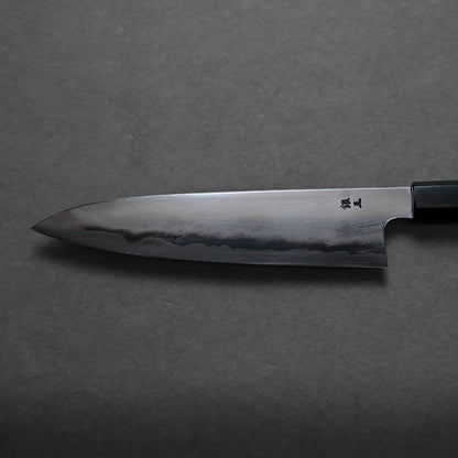 A close-up view of a Japanese chef’s knife with a dark steel blade and a straight-edged handle, lying on a dark, textured surface. The blade features a visible hamon line and displays traditional Japanese craftsmanship.