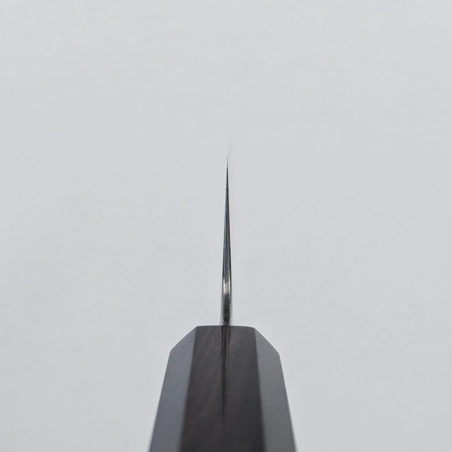 Choil shot of a knife blade displaying a thin, straight edge, positioned vertically against a plain white background for detailed examination.
