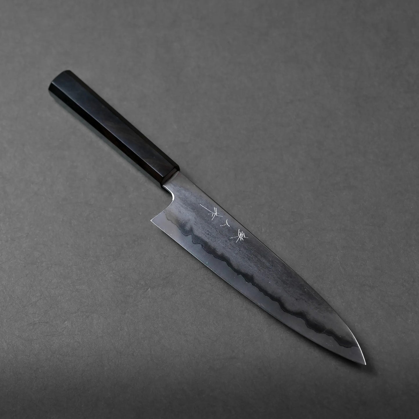 A Japanese chef’s knife with a dark wooden handle and a beautifully patterned steel blade rests diagonally on a gray surface. Intricate Japanese characters are engraved on the blade, highlighting its authentic craftsmanship.