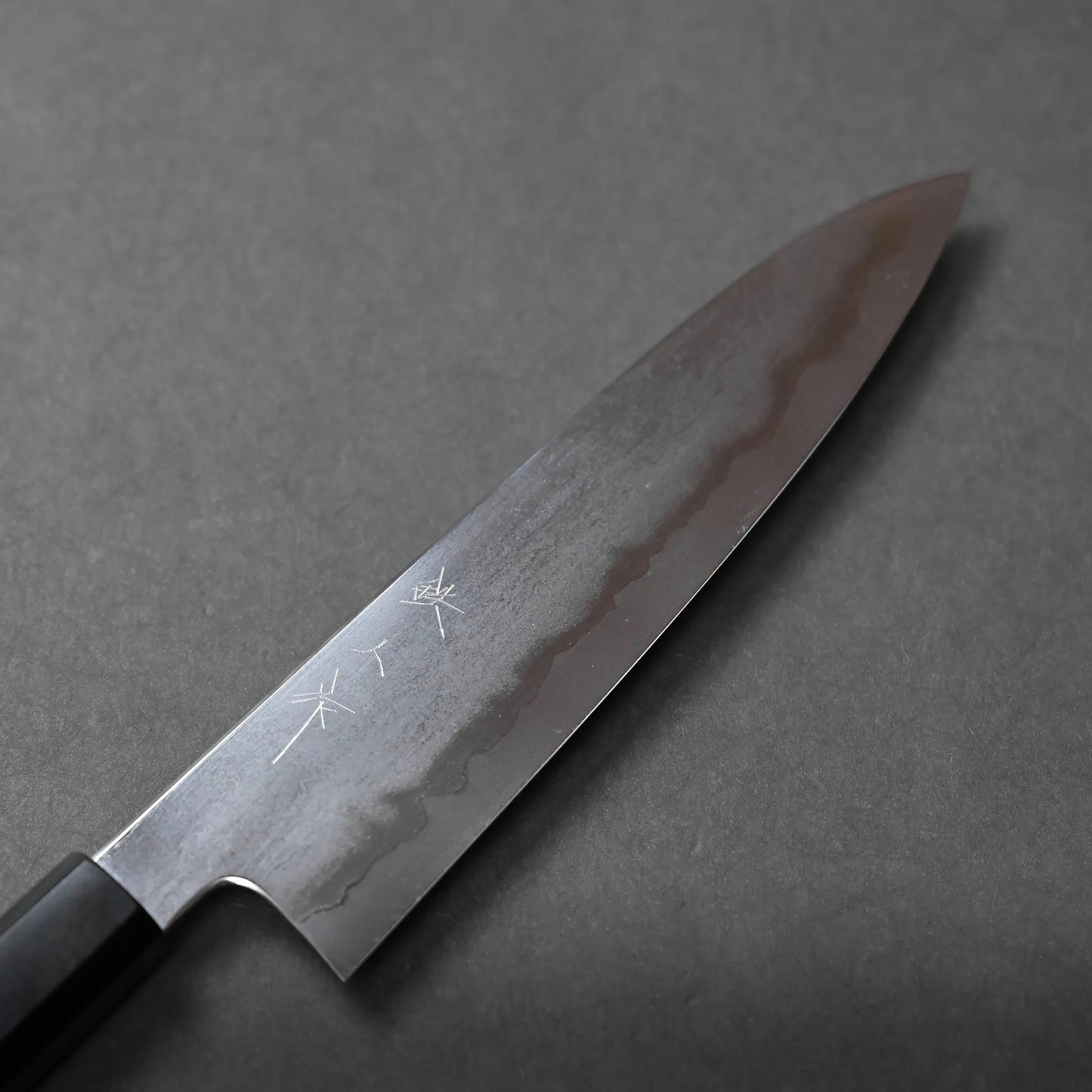 A Japanese chef’s knife with a dark handle and Damascus steel blade rests on a grey surface. The blade displays a wavy pattern and engraved kanji characters, highlighting traditional craftsmanship.