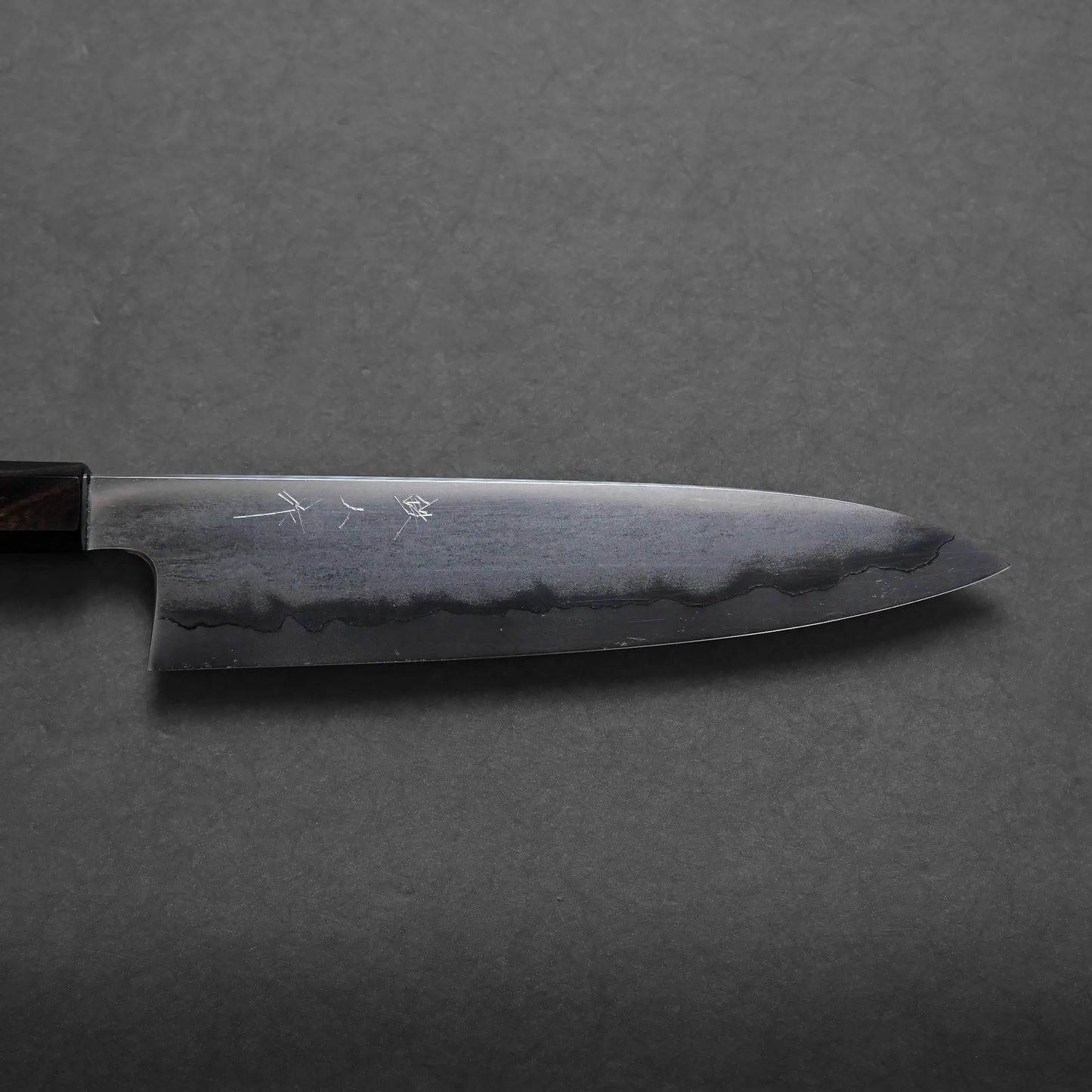 A close-up view of a Japanese chef’s knife with a dark blade, visible wavy hamon line, and a partial wooden handle resting on a dark, textured surface highlights its craftsmanship.