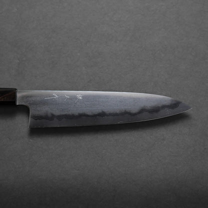 A close-up view of a Japanese chef’s knife with a dark blade, visible wavy hamon line, and a partial wooden handle resting on a dark, textured surface highlights its craftsmanship.