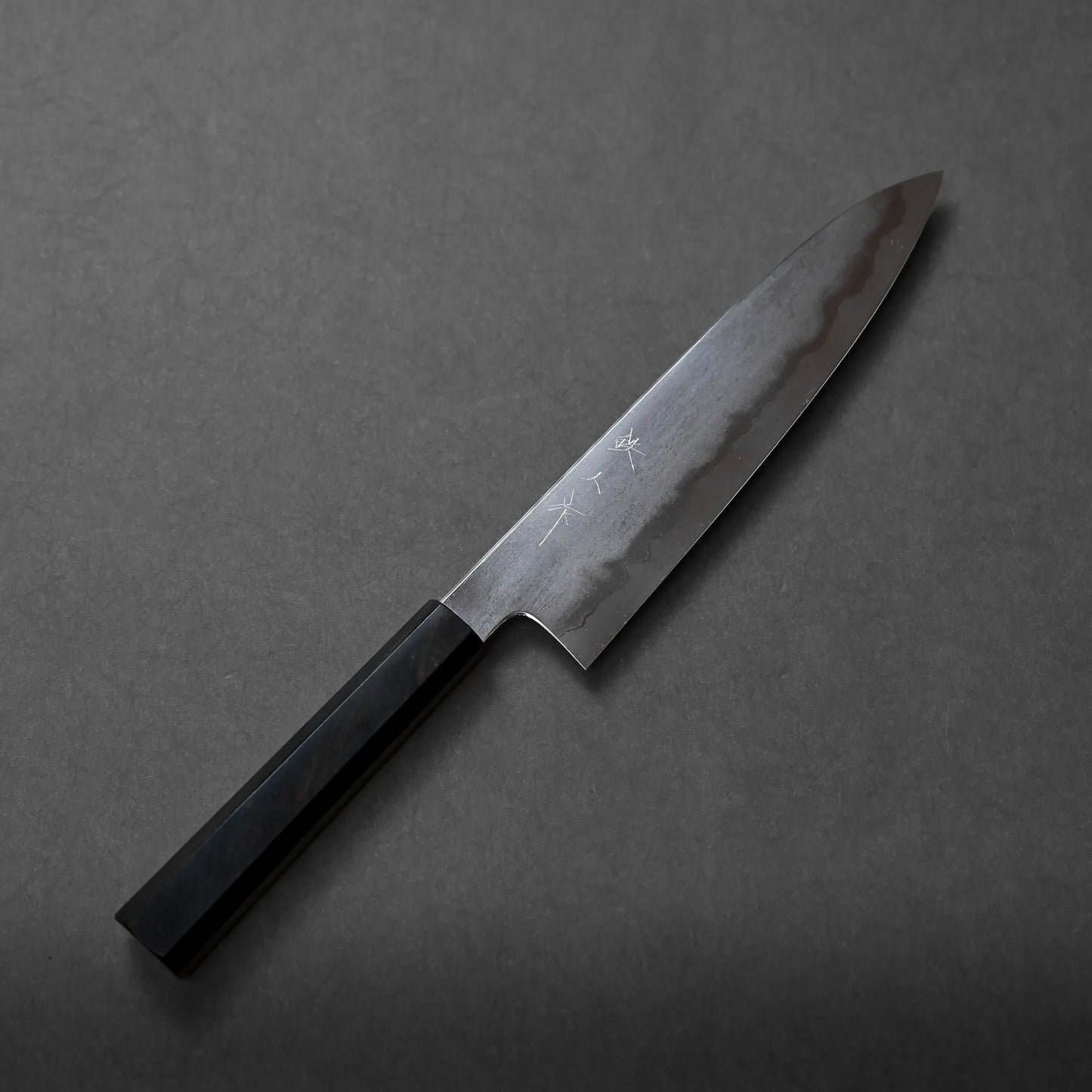 Tetsujin tanryusen ginsan gyuto 210mm chef’s knife with a dark wooden handle and a polished, engraved blade rests on a plain dark gray surface, reflecting artisanal craftsmanship.