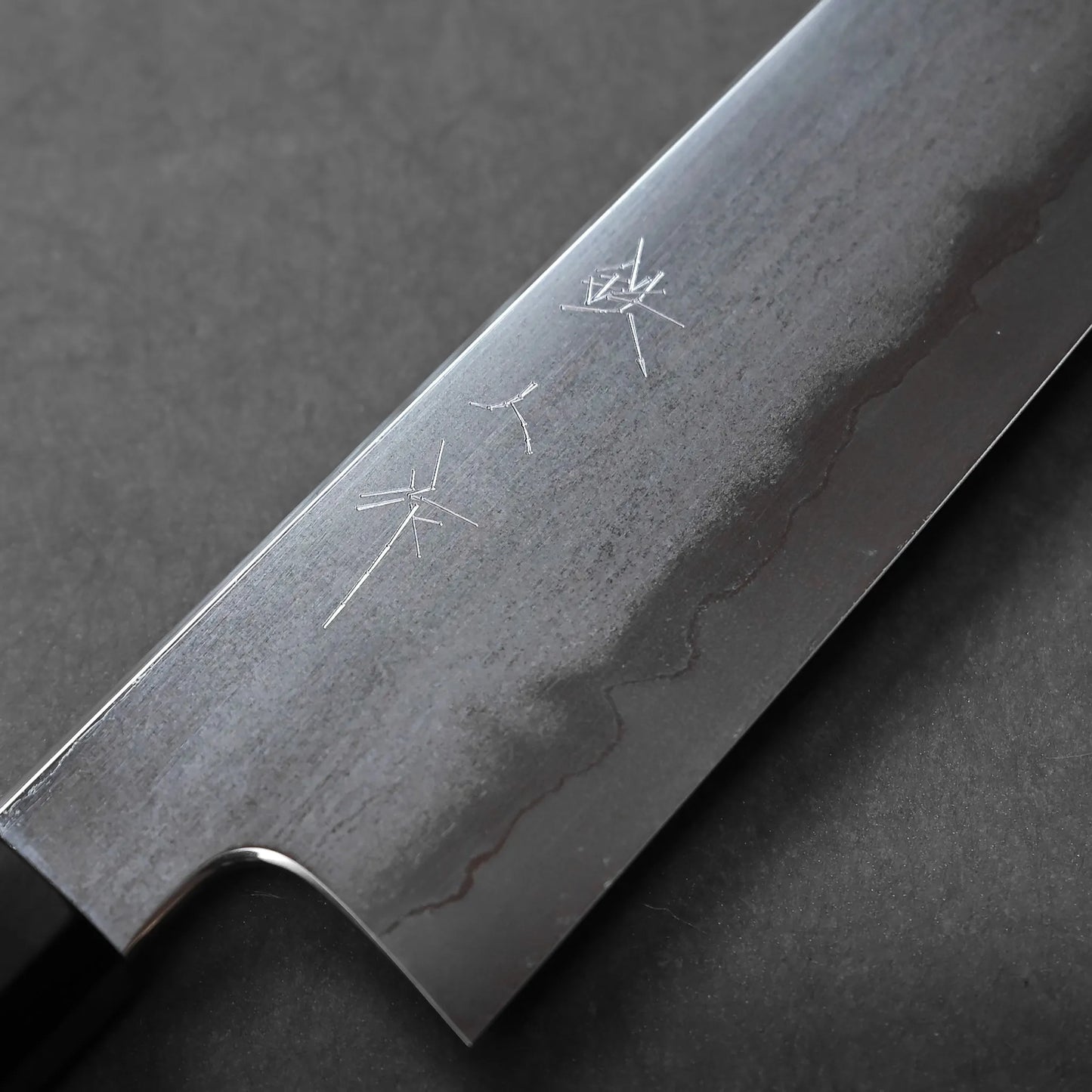 Close-up of a Japanese kitchen knife blade featuring a wavy Damascus pattern and engraved kanji characters, set against a dark, textured background.
