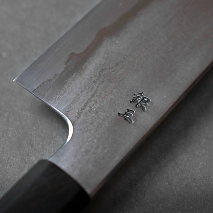 A close-up of a Japanese kitchen knife with kanji characters engraved on the blade, resting on a dark, smooth surface, showcases traditional craftsmanship.