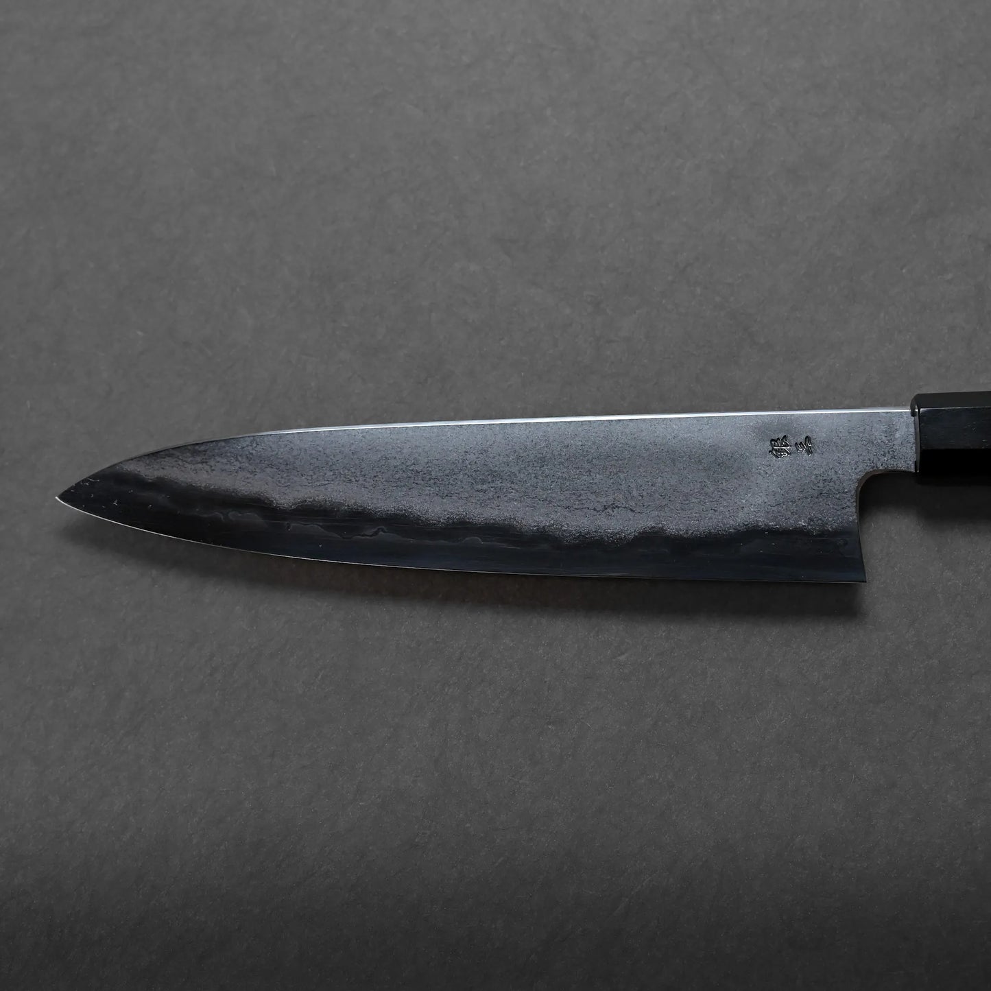 A close-up of a Japanese-style chef’s knife with a hammered, black Damascus blade and a straight edge, positioned on a plain, dark gray surface.