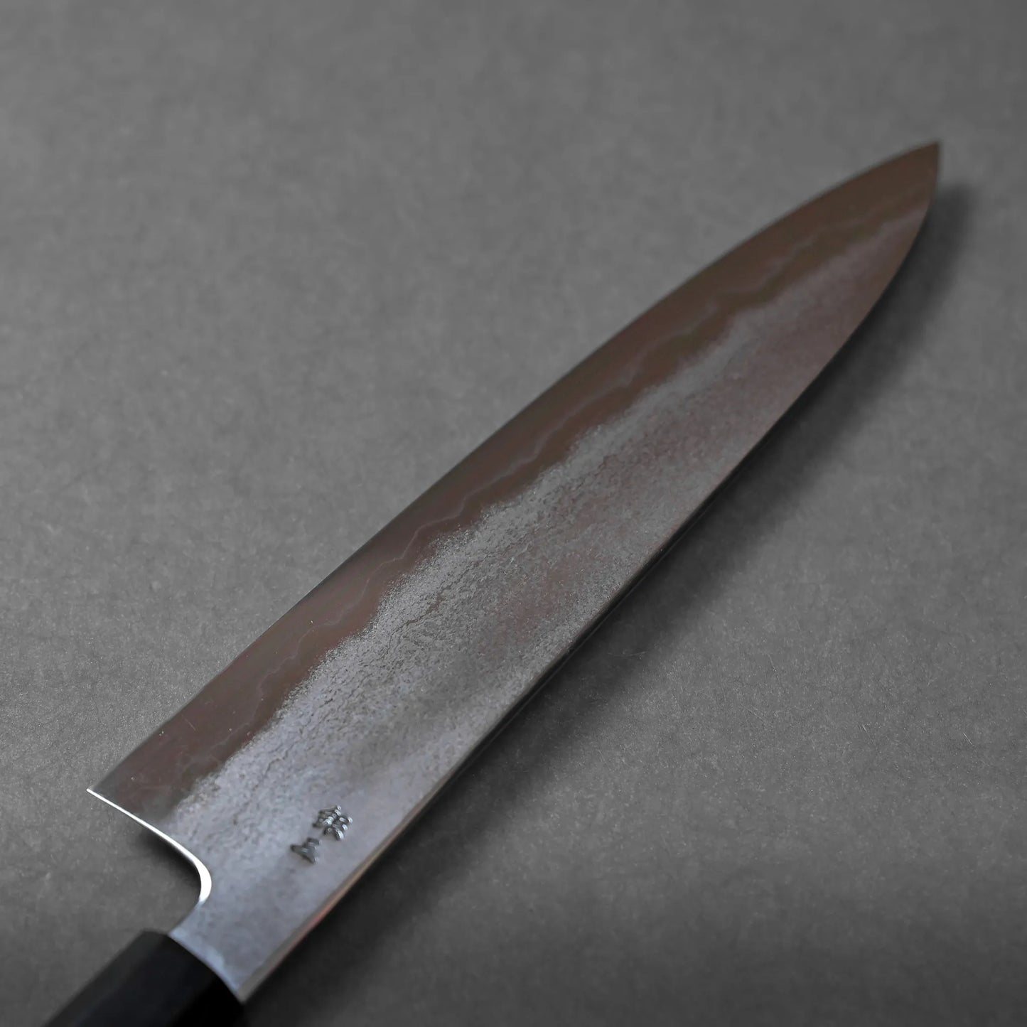 A close-up of a Japanese chef’s knife featuring a beautiful wavy pattern on the blade, photographed on a plain gray background.