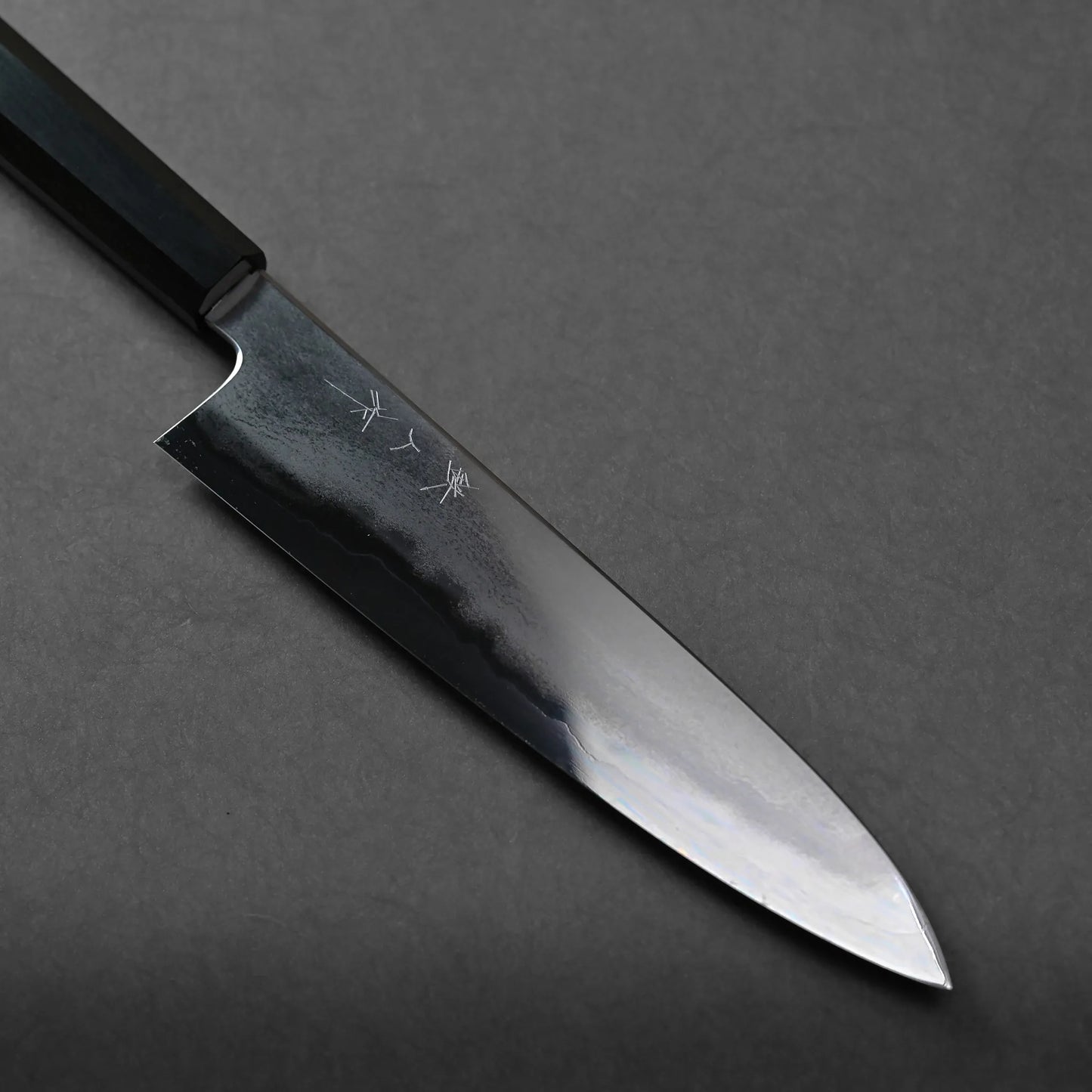 A Japanese chef’s knife with a dark handle and a polished, partially reflective blade rests on a black surface. The blade features subtle wave patterns, engraved Japanese characters, and exceptional craftsmanship.