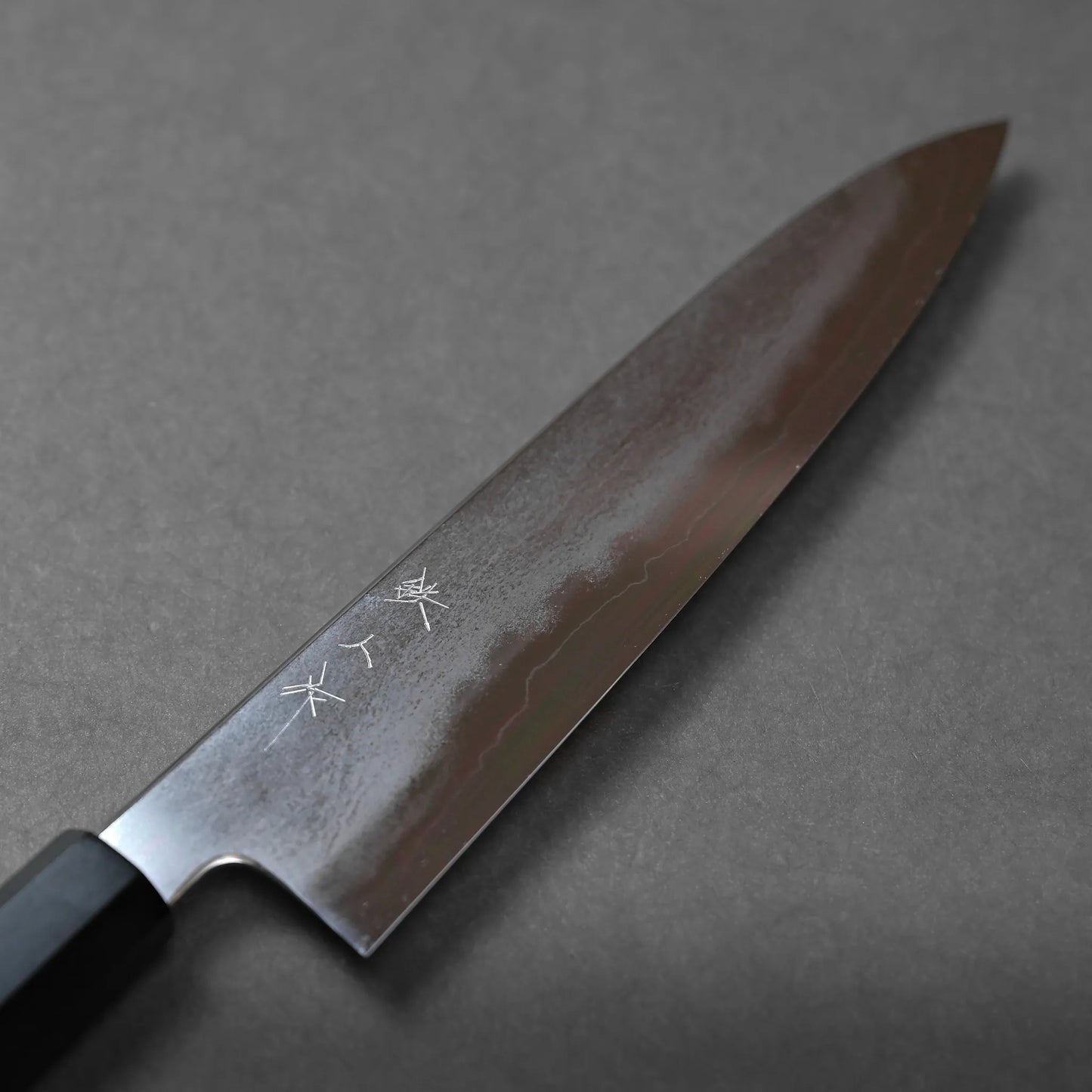 A close-up of a Japanese chef’s knife with a dark wooden handle and a polished blade featuring engraved Japanese characters, set against a plain gray background.