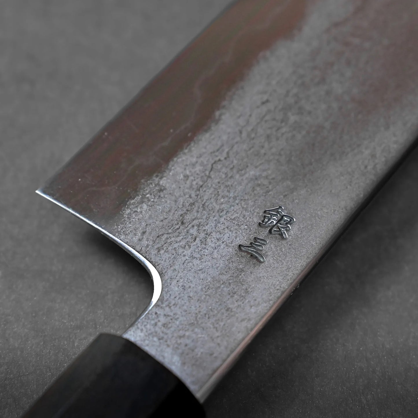 A close-up of a Japanese kitchen knife blade displays wavy patterns and engraved Japanese characters near the edge, with a Damascus steel finish set against a neutral grey background.