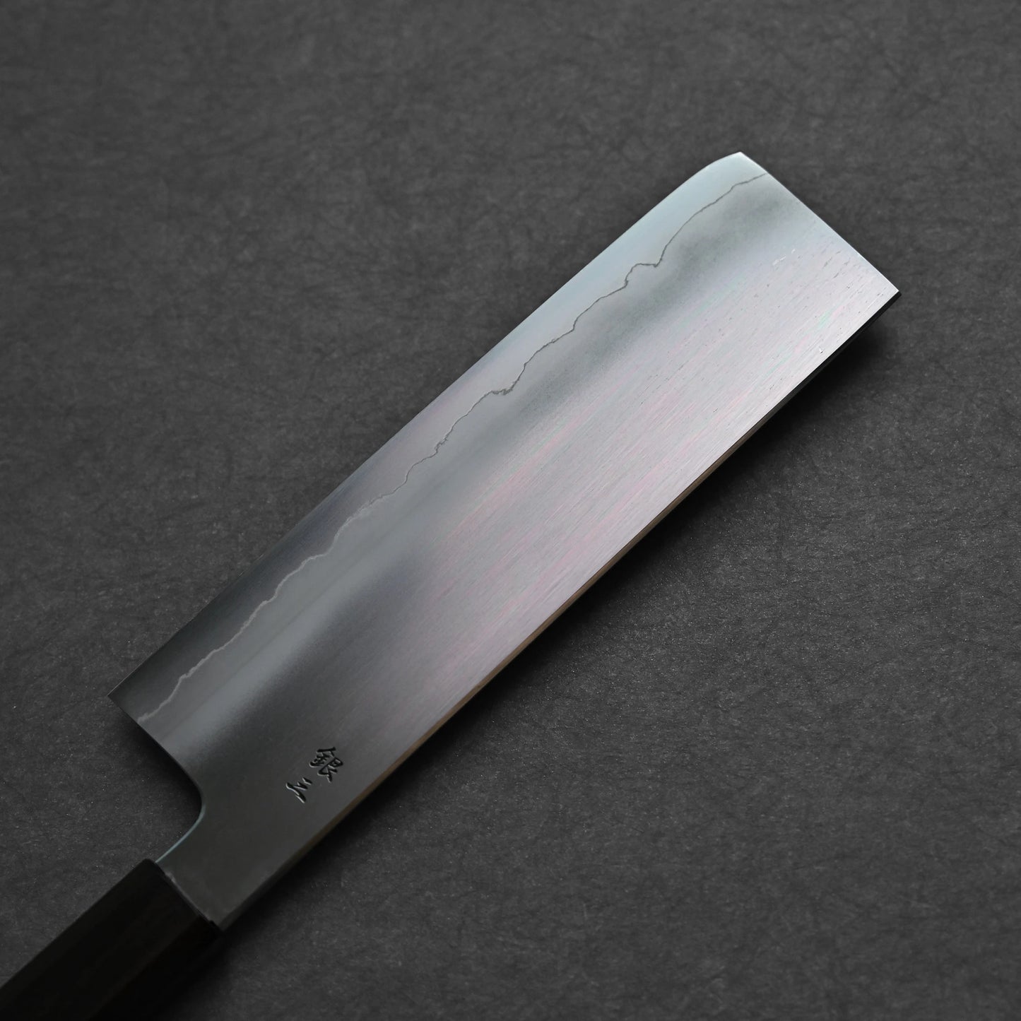 Nakiri knife photographed from the reverse side, showing a smooth steel surface with three vertically engraved kanji characters near the spine; close-up angle emphasizes finish clarity and traditional craftsmanship.
