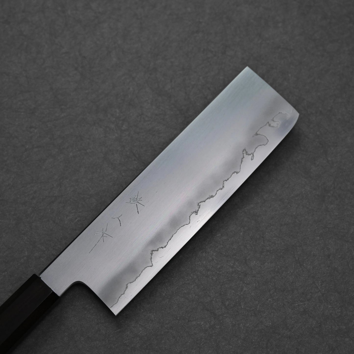 Nakiri knife featuring a zoomed-in view of the rectangular blade with vertically engraved kanji near the spine; paired with a dark octagonal handle and photographed against a textured surface to emphasize steel finish and engraving detail.