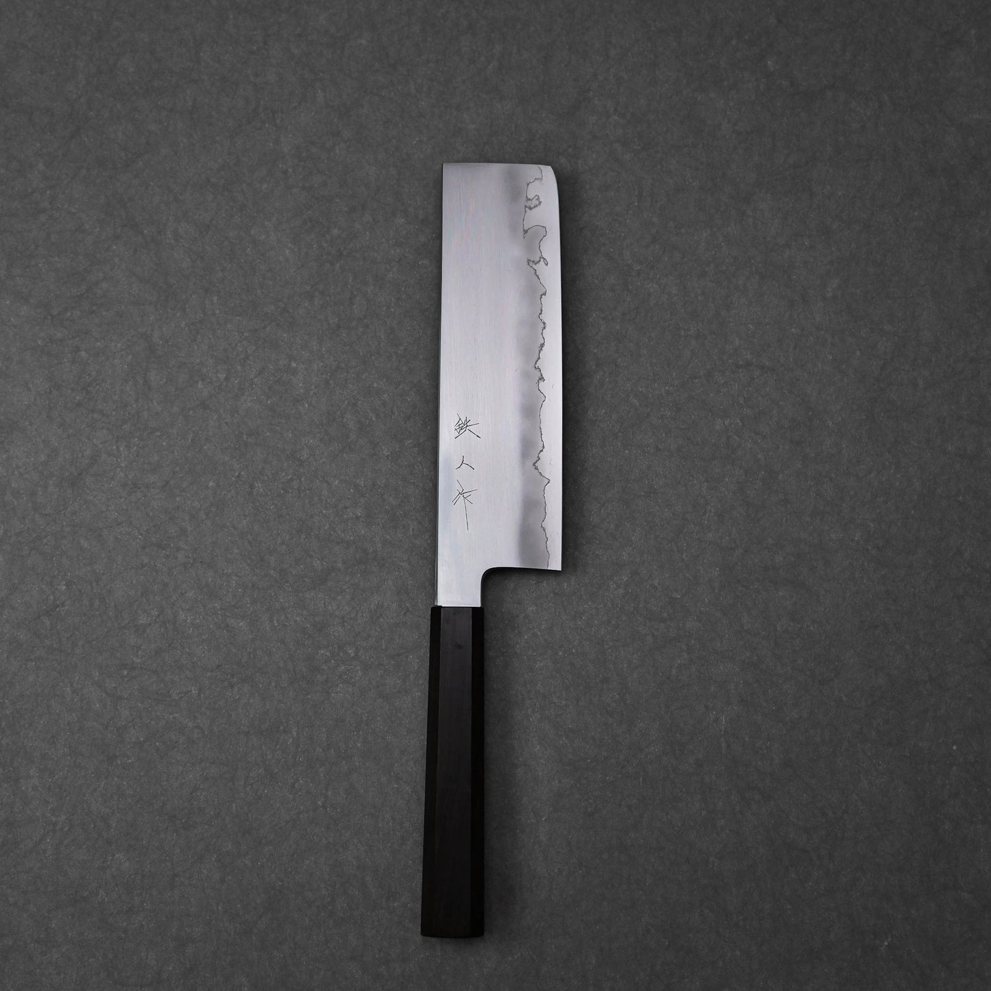 Nakiri knife showing the full blade profile with three vertically engraved kanji characters near the spine; photographed to highlight the polished steel surface and overall craftsmanship.