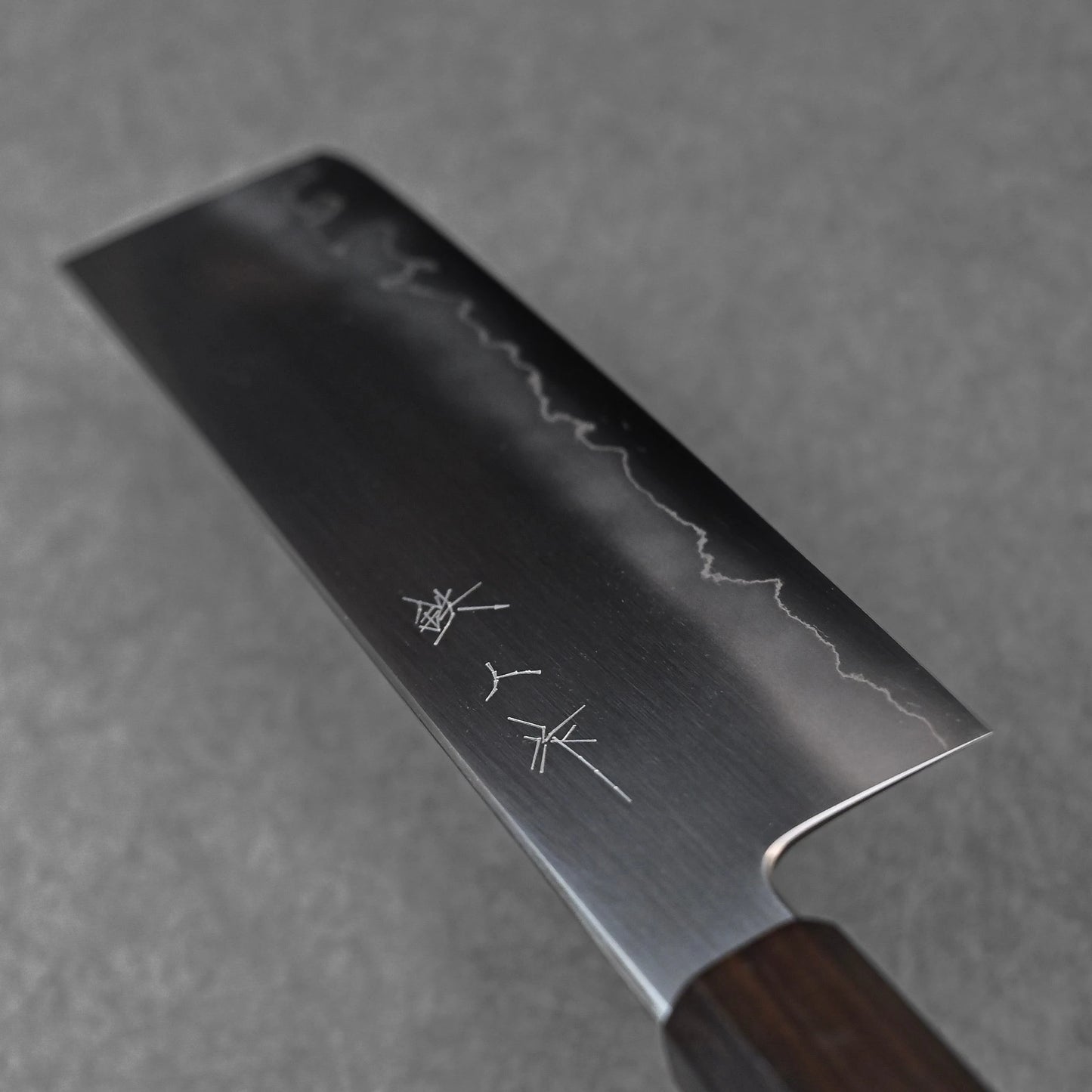 Nakiri knife featuring three vertically engraved kanji characters near the spine; photographed close-up to highlight the polished steel surface and craftsmanship detail.