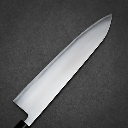 A close-up of a sharp kasumi gyuto kitchen knife with a reflective blade and black handle, placed on a dark gray textured surface.