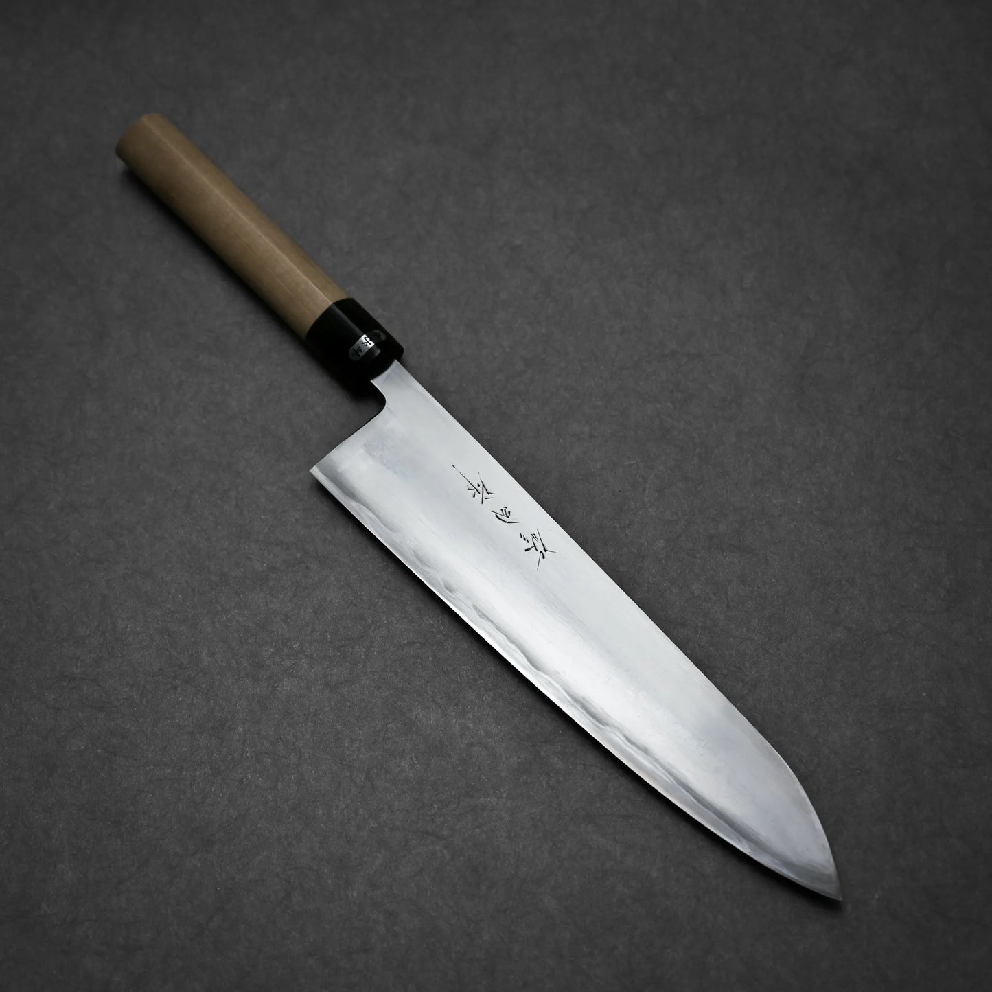 A Sanjo gyuto chef’s knife with a wooden handle and a shiny, sharp blade featuring engraved Japanese characters rests on a dark gray surface.