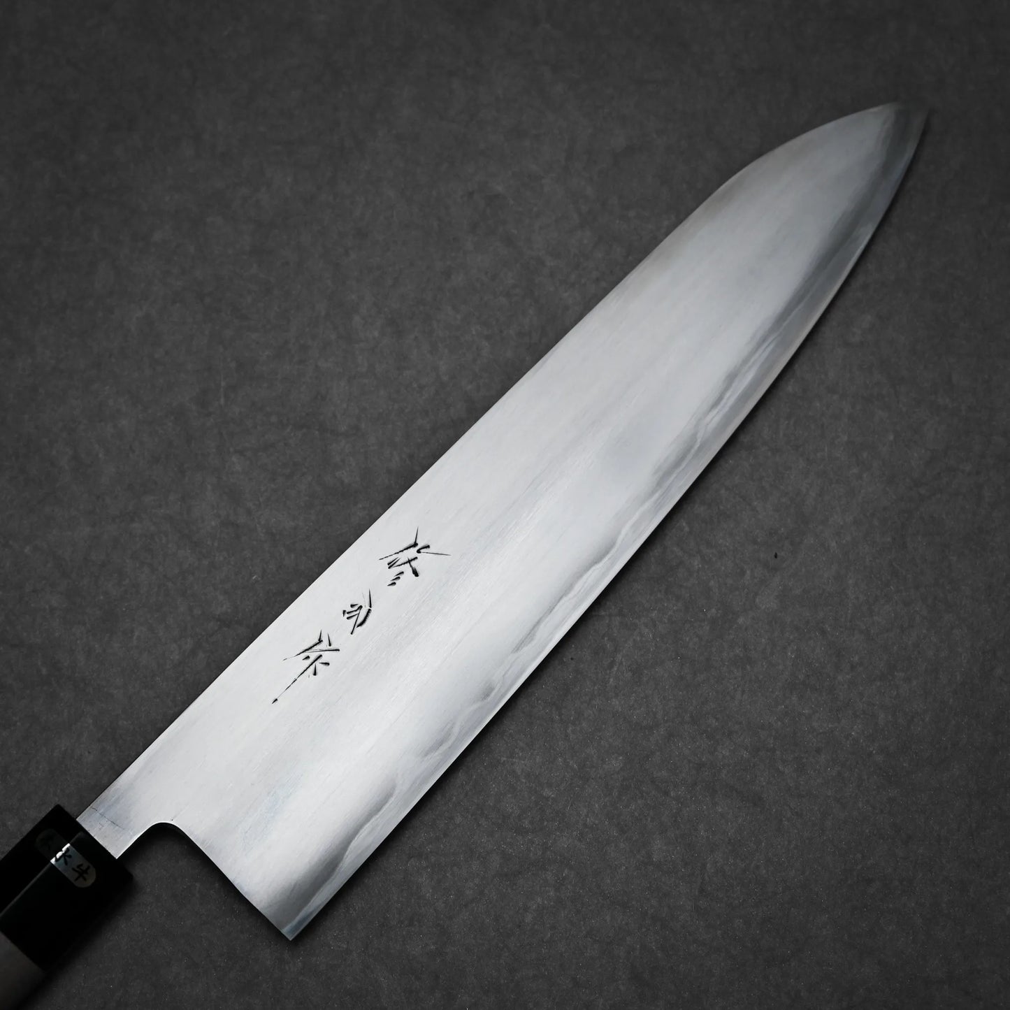 A close-up of a kasumi gyuto Japanese kitchen knife with a polished steel blade and black handle, featuring engraved Japanese characters on the blade, placed on a dark, textured background.