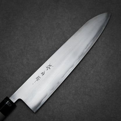 A close-up of a kasumi gyuto Japanese kitchen knife with a polished steel blade and black handle, featuring engraved Japanese characters on the blade, placed on a dark, textured background.