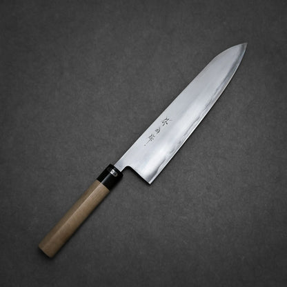 A Toyama aogami#2 gyuto 270mm with a wooden handle and a sharp, shiny blade rests on a dark gray surface. The blade, featuring Japanese characters engraved near the handle, showcases traditional craftsmanship.