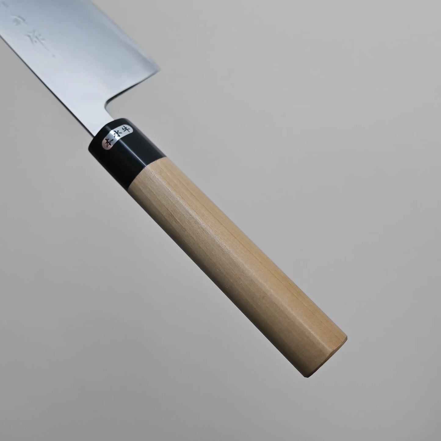 A close-up of the handle and part of the blade of a Sanjo gyuto kitchen knife with a light wood handle and metal blade, photographed against a plain gray background.