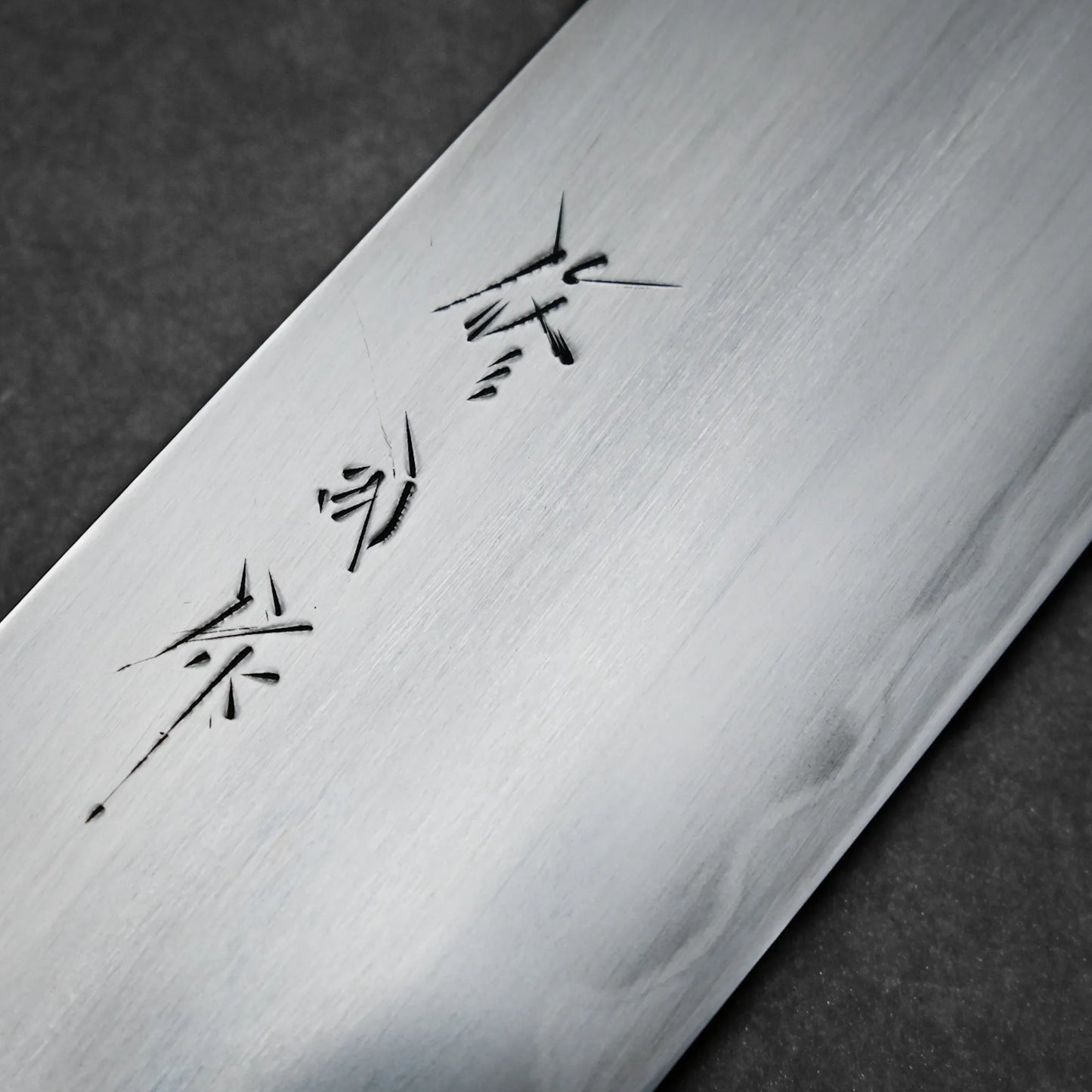 Close-up of a metallic kasumi gyuto blade featuring engraved Japanese characters, set against a dark, textured background.