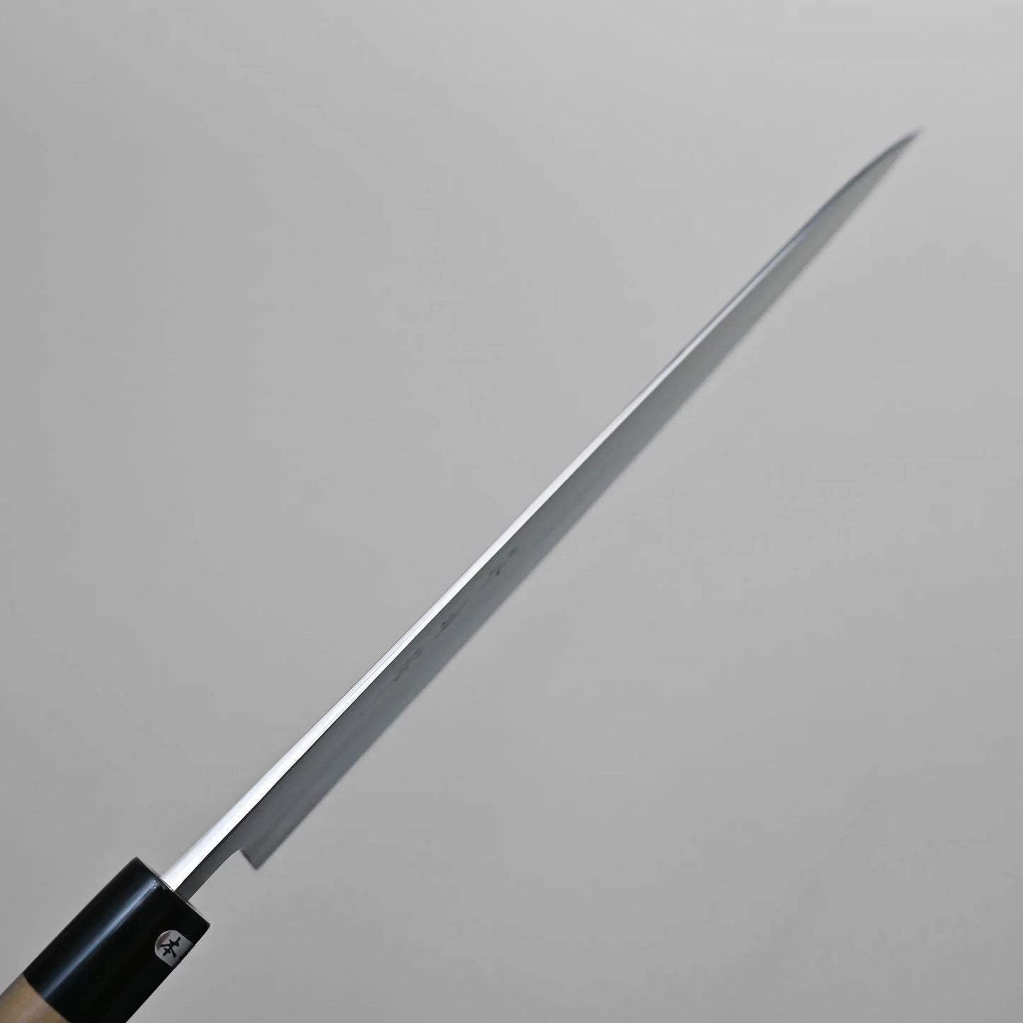 Spine shot of a thin, long-bladed Sanjo gyuto kitchen knife held at an angle against a plain, light gray background. The blade appears sharp and tapers to a pointed tip.