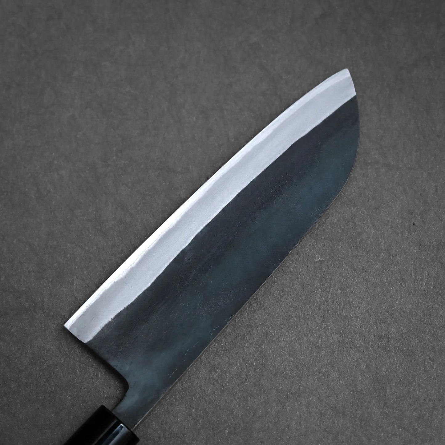A close-up view of a kurouchi santoku with a dark blade and visible lighter edge, resting on a textured grey surface.