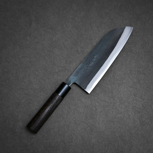 A Toyama kurouchi aogami#2 santoku 180mm with a dark wooden handle and a broad, sharp blade featuring engraved characters, placed on a textured dark gray surface.