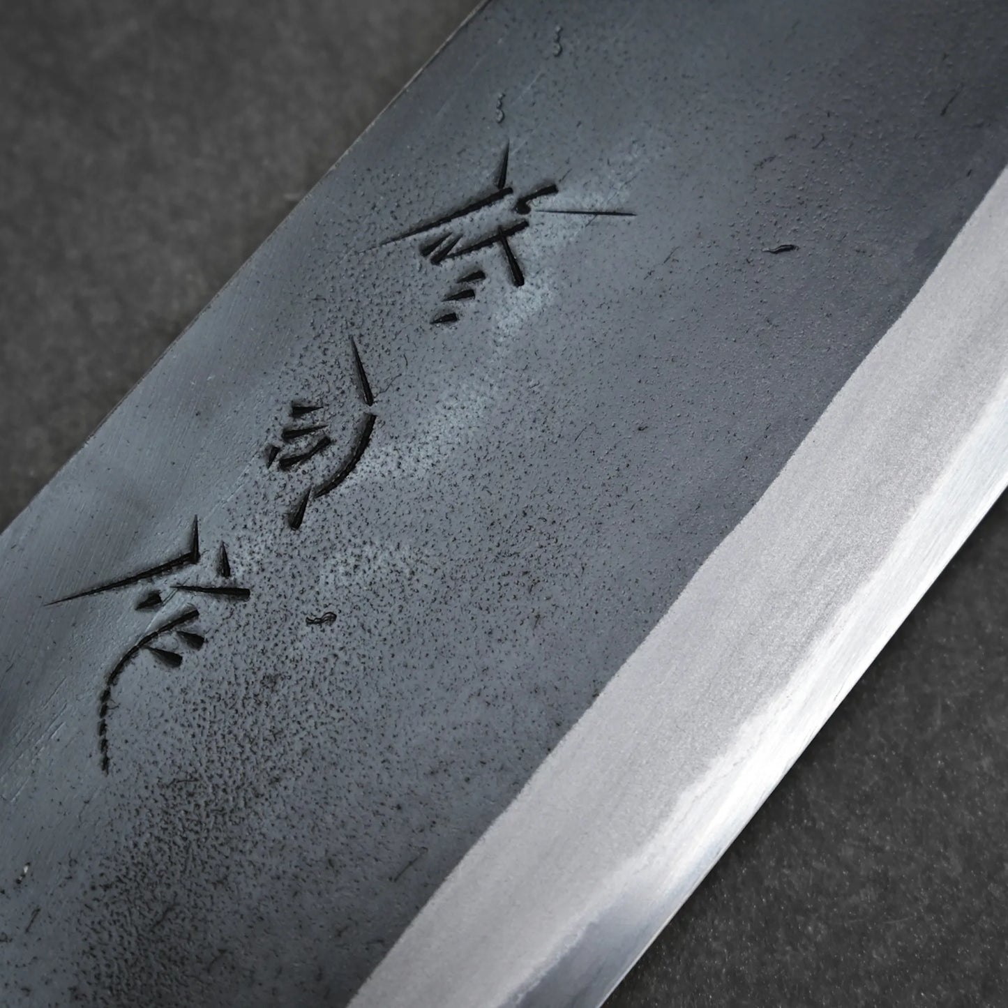 Close-up of a kurouchi santoku blade with a matte finish and Japanese characters engraved on the surface, placed on a dark background.