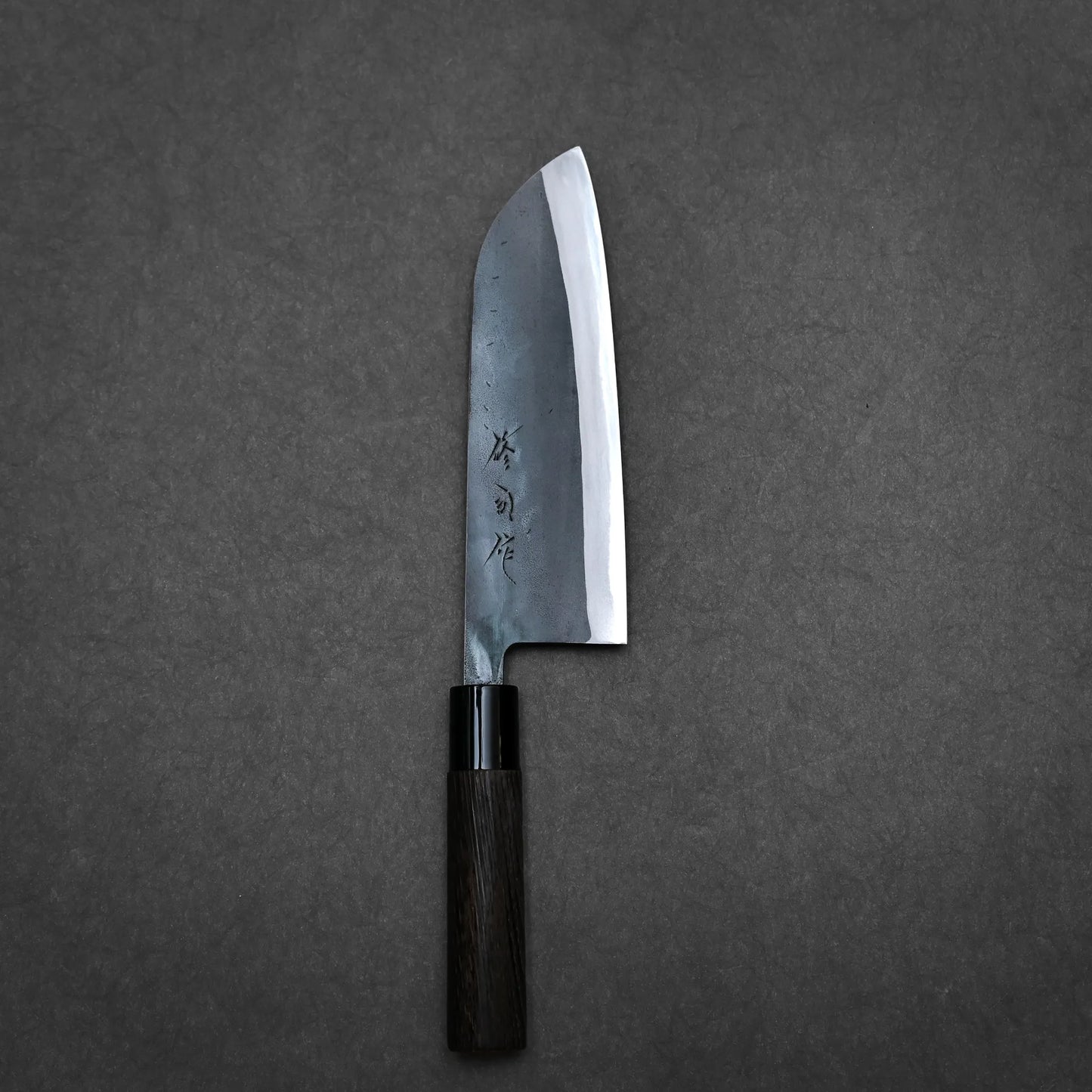 A Japanese-style kurouchi santoku kitchen knife with a black handle and a wide, straight-edged stainless steel blade featuring engraved Japanese characters, placed on a dark gray surface.