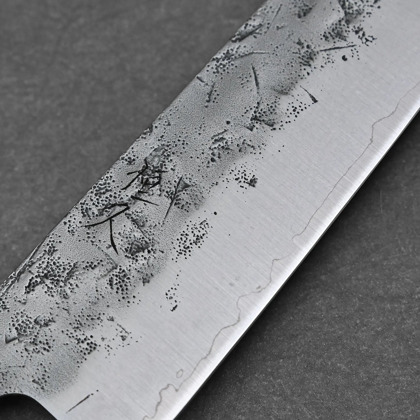 A close-up of the Tsunehisa nashiji SLD bunka 170mm blade highlights intricate nashiji patterns and fine lines on the SLD steel, with visible dark etching, set against a plain dark background.