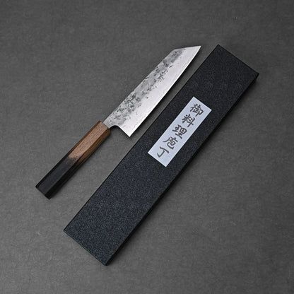 The Tsunehisa nashiji SLD bunka 170mm—a Japanese kitchen knife with a wooden handle—rests beside a black textured box with a white Japanese label, all arranged on a dark gray surface.