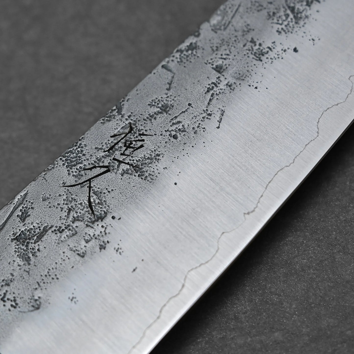 Close-up of the Tsunehisa nashiji SLD gyuto 210mm blade, showing its textured nashiji finish and engraved markings, with a subtle wavy line near the edge against a dark background.