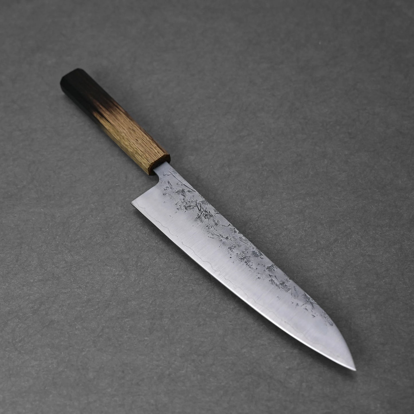 The Tsunehisa nashiji SLD gyuto 210mm features a wide, slightly curved, textured mottled blade made from SLD steel and a wooden handle with a dark-to-light gradient finish, shown resting on a gray surface.