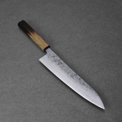 The Tsunehisa nashiji SLD gyuto 210mm features a wide, slightly curved, textured mottled blade made from SLD steel and a wooden handle with a dark-to-light gradient finish, shown resting on a gray surface.