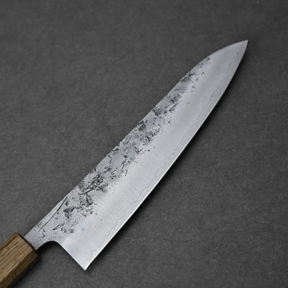 A close-up of the Tsunehisa nashiji SLD gyuto 210mm, featuring a wooden handle and a partially textured SLD steel blade with a nashiji (pear skin) finish, lying on a dark grey surface.