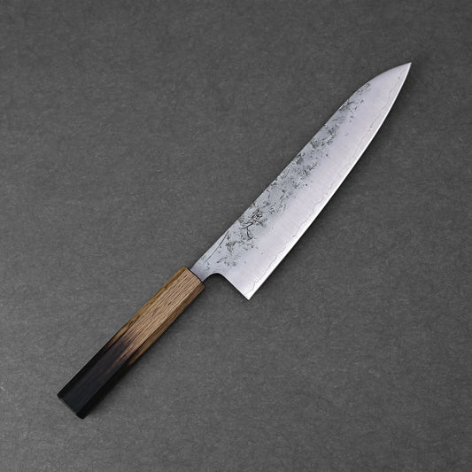 The Tsunehisa nashiji SLD gyuto 210mm, featuring a patterned SLD steel blade and wooden handle, rests on a dark gray textured surface.