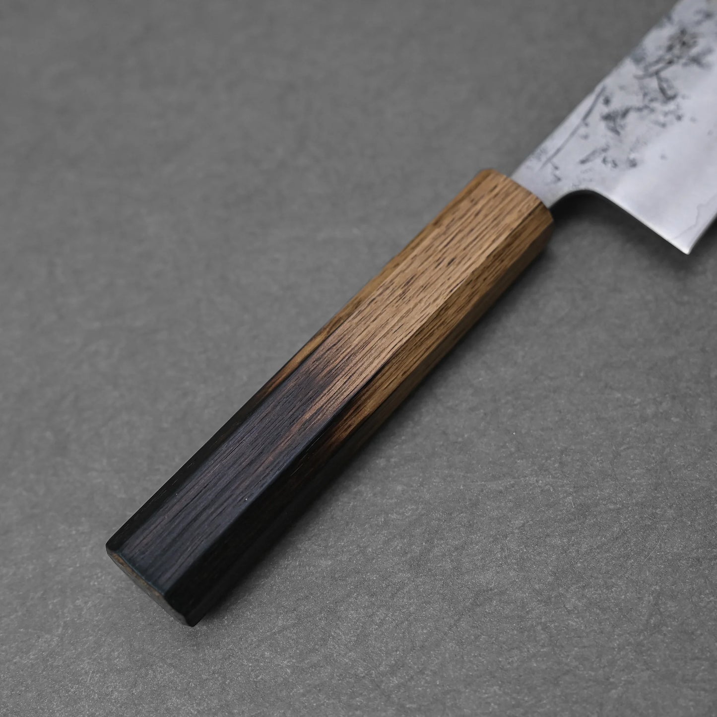 A close-up of the Tsunehisa nashiji SLD gyuto 210mm, featuring a wooden handle and a partially visible SLD steel blade, lying on a gray textured surface.