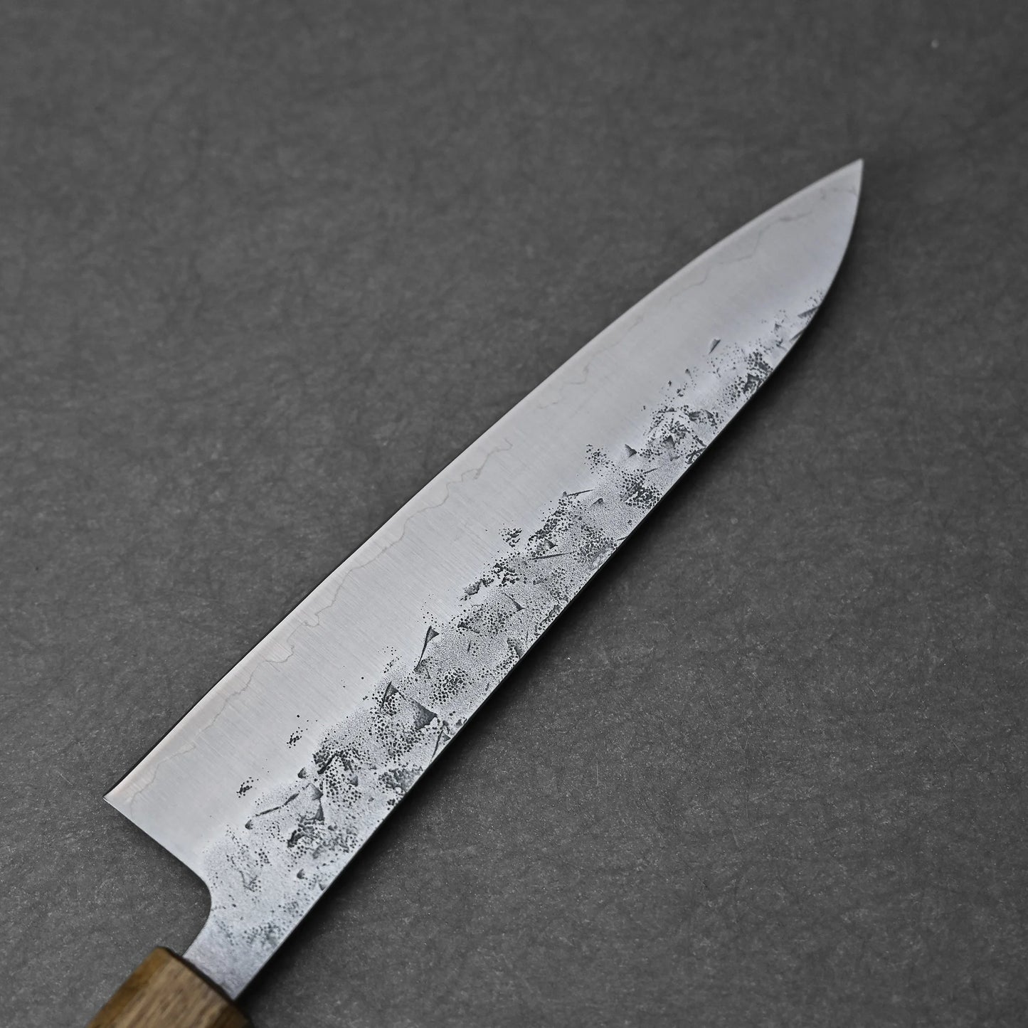 A close-up of the Tsunehisa nashiji SLD gyuto 210mm, featuring a wooden handle and a patterned SLD steel blade, set against a gray stone background.