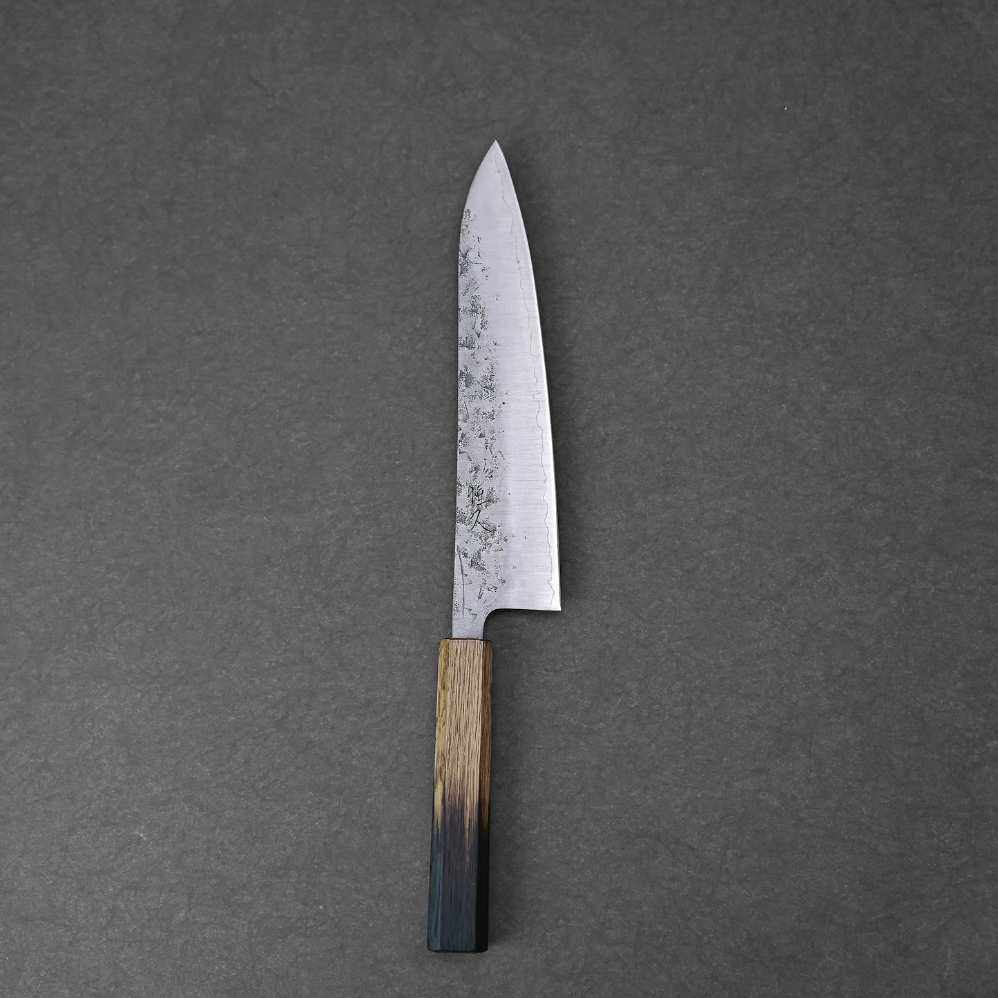 A Tsunehisa nashiji SLD gyuto 210mm with a long, narrow SLD steel blade and a two-tone wooden handle rests on a plain, dark gray surface.