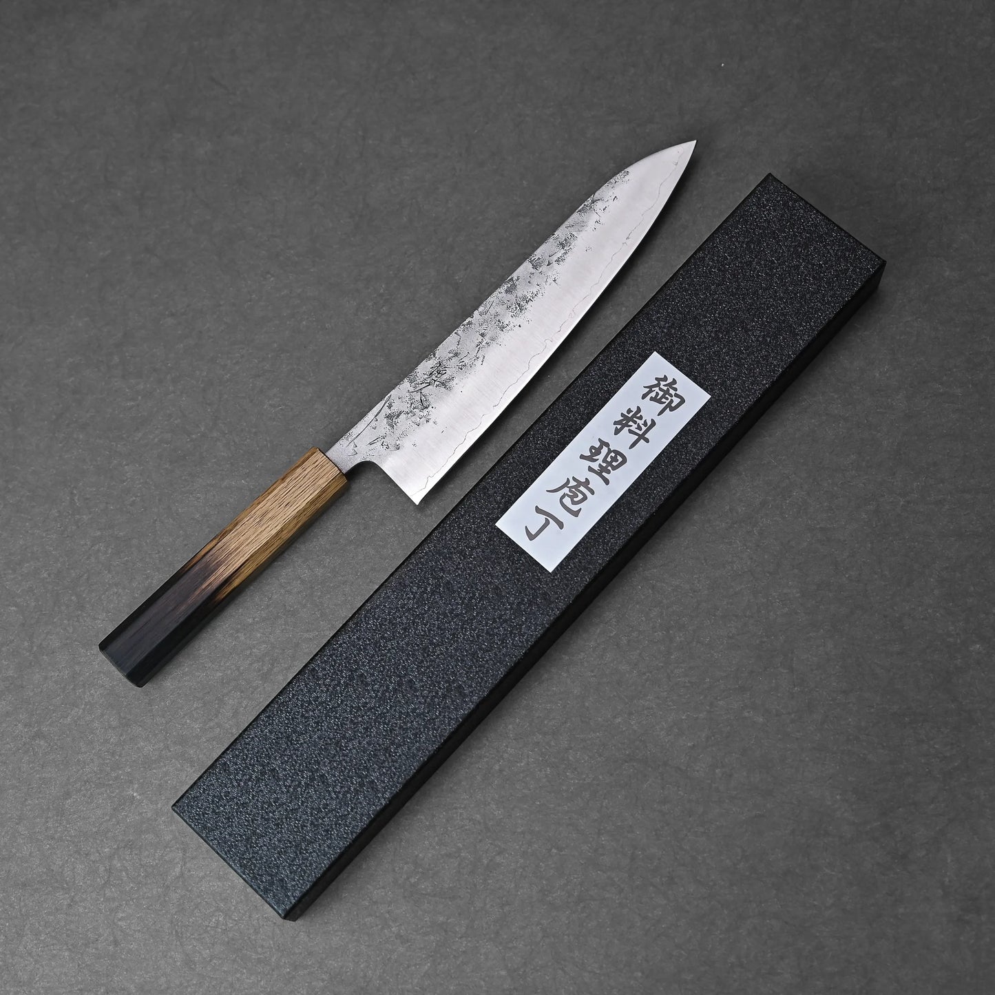 A Tsunehisa nashiji SLD gyuto 210mm with a wooden handle rests beside a rectangular black box bearing a white label with Japanese characters, all placed on a dark gray textured surface.