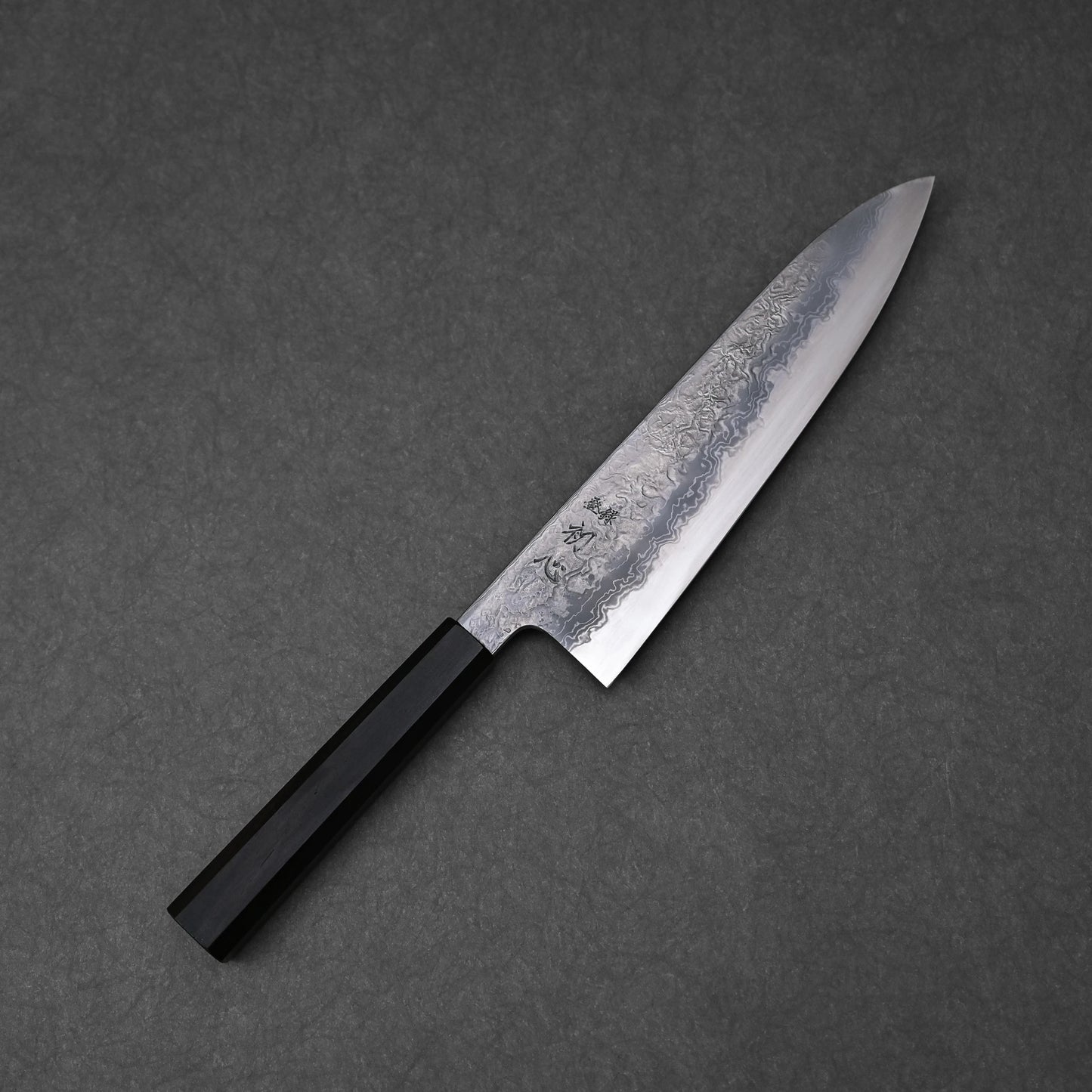 Japanese chef’s knife with a damascus steel blade featuring layered forge patterns and vertically engraved kanji near the handle; paired with a black octagonal handle and photographed on a dark textured surface.