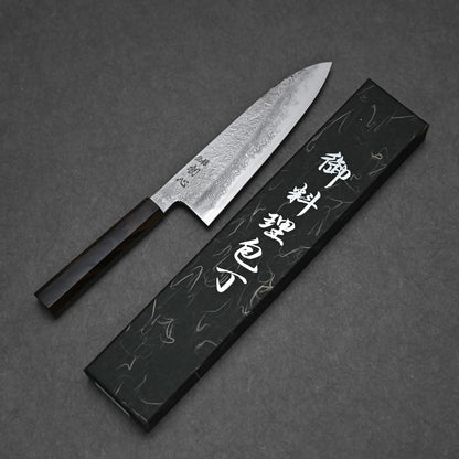 The Yoshihide Masuda aogami#1 damascus santoku 180mm, featuring a damascus finish and dark wooden handle, rests beside a black box with white Japanese lettering on a dark surface.