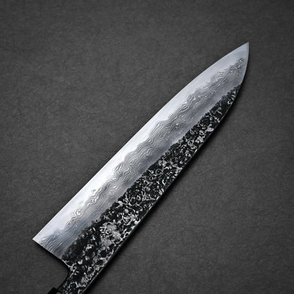 A close-up of a kurouchi damascus gyuto blade with a distinct wavy pattern and textured black area near the edge, set against a dark grey background.