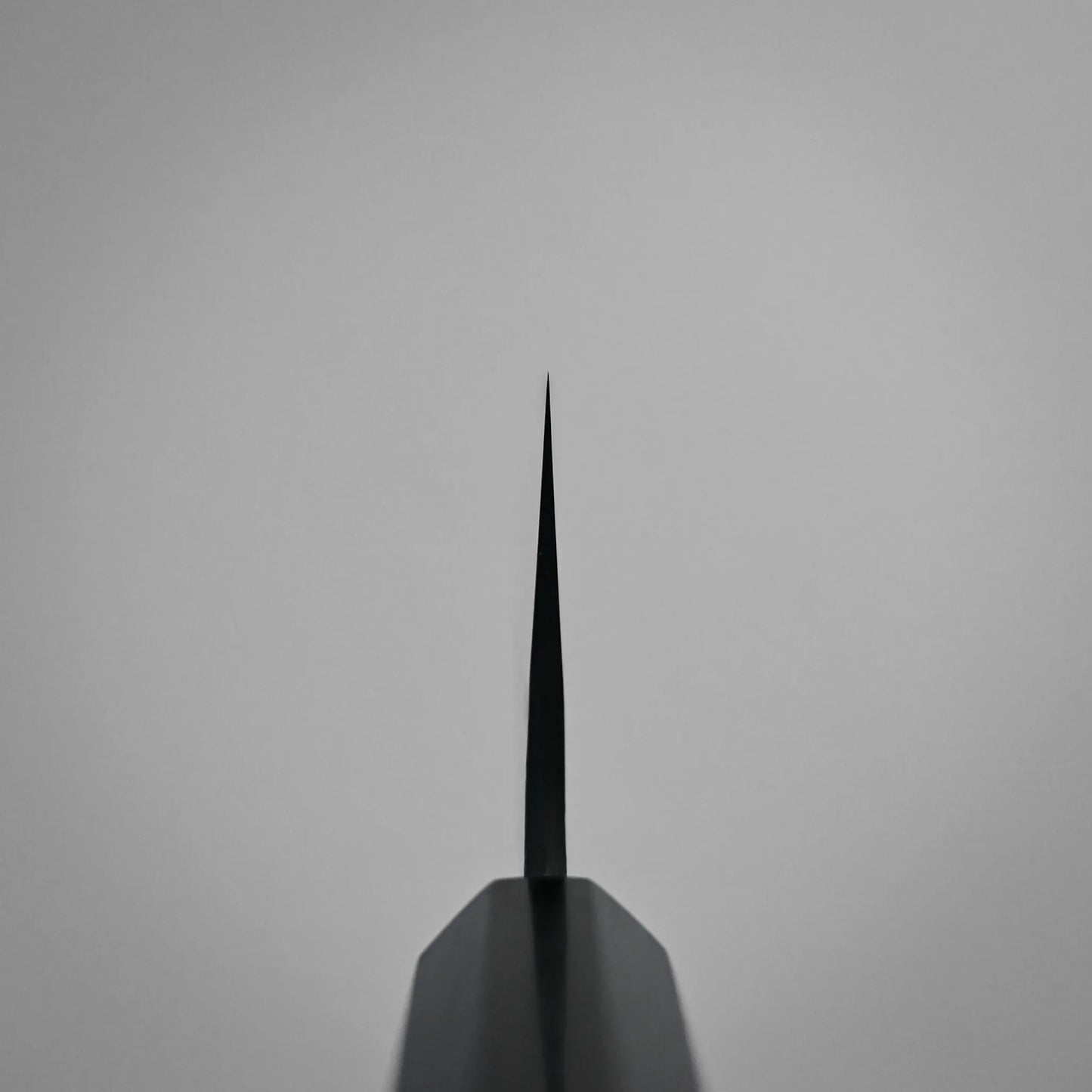 Choil shot of a tall gyuto knife blade, pointed directly upward against a plain, light gray background. The image emphasizes the thinness and precision of the blade’s edge.