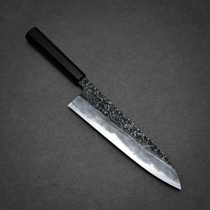 A kurouchi damascus gyuto with a dark handle and a patterned steel blade, featuring a textured upper section and polished lower section, is placed on a dark gray surface.