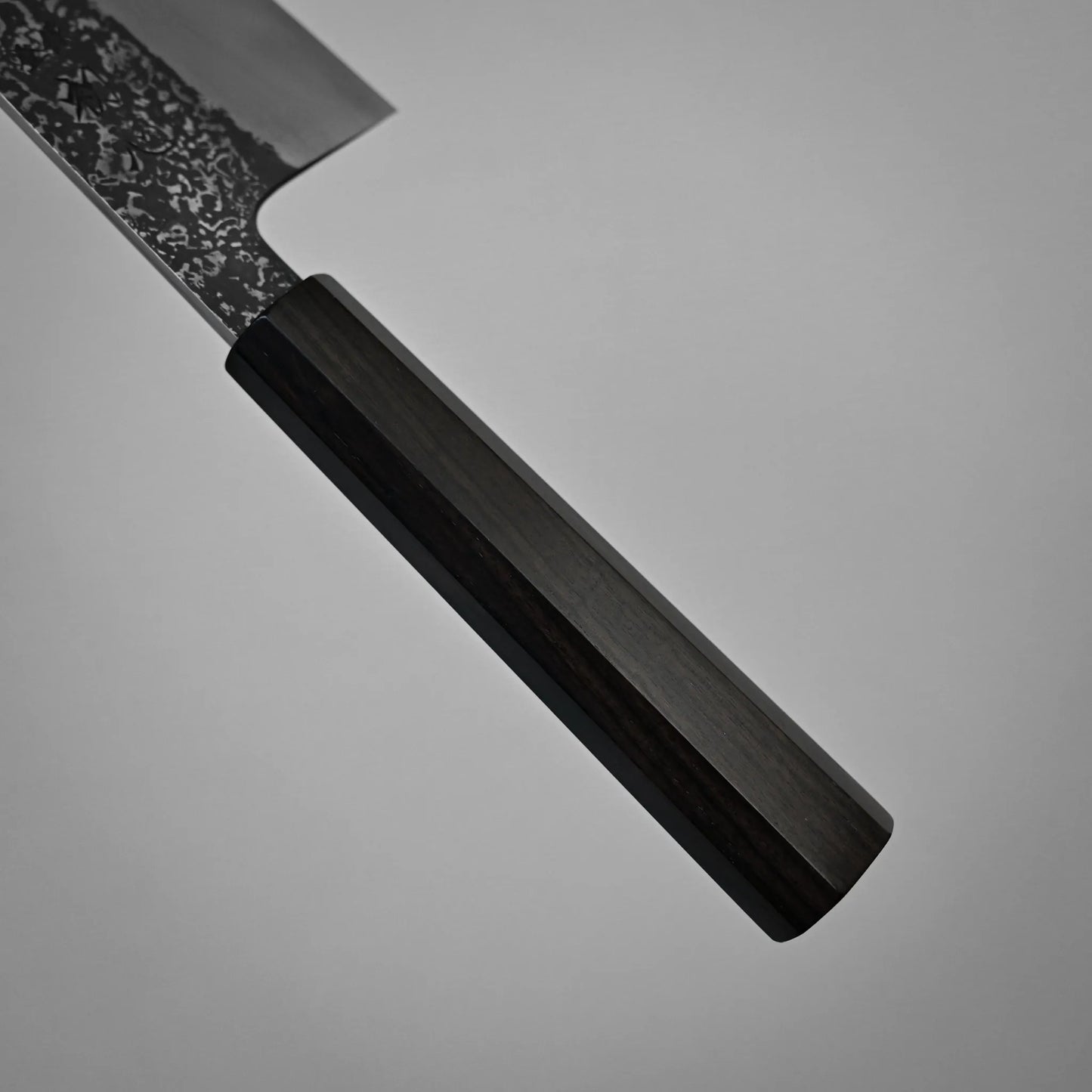 A close-up of a kurouchi damascus gyuto reveals a dark wooden handle and part of its textured metal blade against a plain grey background.